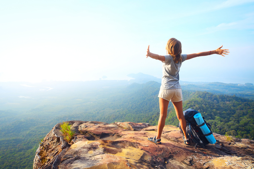 young happy woman with backpack standing on a rock with raised hands and looking to a valley below